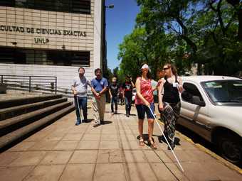  tres personas con los ojos vendados, con baston blanco y acompañante caminan por las veredas de la Facultad 